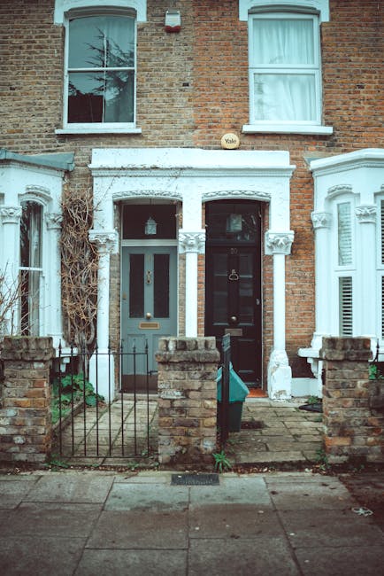 A row of two adjoining residential terraced houses with brick facades and white decorative trim around the doorways and windows. The ground-floor windows and doors feature glazed panes; one door is painted light blue, and the other is black, both with metallic handles and letterboxes. The small front yards are enclosed by low brick walls and metal fences, with paved pathways leading to each entrance. The upper-story windows are framed with white casings, and the left-side window shows a reflected tree branch. The scene is illuminated by natural daylight, emphasizing the clean appearance of the brickwork and the tidy, well-maintained appearance of the facades. This image represents the type of property serviced by Cleaners Finsbury Park, showcasing traditional residential architecture typical of the area near Manor House N4 flats, suitable for deep cleaning and surface sanitisation as part of end of tenancy cleaning services.