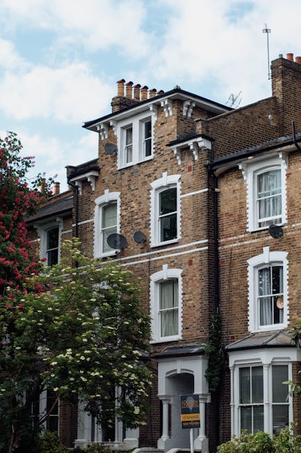 A multi-story residential building constructed with red brick featuring white window frames and decorative trim. The building has multiple satellite dishes attached to its exterior walls and a pitched roof with several chimneys. Surrounding trees with green foliage partially obscure the lower floors, and the sky above is partly cloudy with patches of blue. The scene showcases an urban residential setting, with the building likely located near Manor House N4 flats in Finsbury Park, suggesting a typical London Victorian terrace. The overall appearance is clean and well-maintained, reflecting the quality of the property in a historic neighborhood, as seen from the detailed brickwork and classic architectural features.