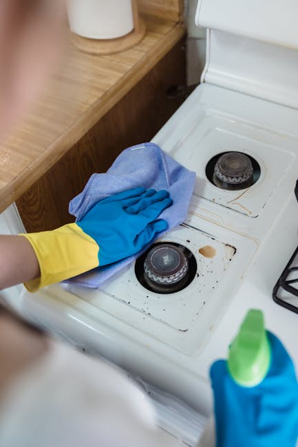 A cleaning professional wearing yellow and blue rubber gloves is using a blue cloth to wipe around the burners on a white ceramic gas hob in a domestic kitchen, with visible blackened stains and grease around the burners indicating prior use. The stove is positioned against a wooden countertop, and a green spray bottle is held in the other hand, suggesting ongoing surface cleaning and degreasing. The lighting is bright, highlighting the cleanliness process as part of a detailed deep cleaning or sanitisation. The image reflects the meticulous maintenance services provided by Cleaners Finsbury Park, specialising in surface cleaning and domestic oven hygiene.