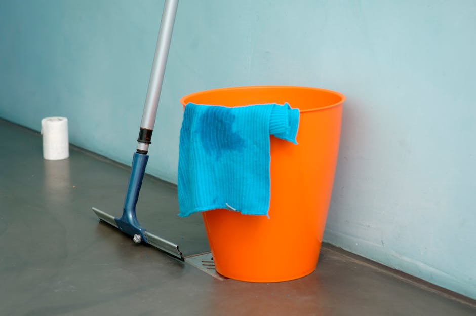 A person wearing light green rubber gloves is performing surface cleaning on a wooden shelf in a room with neutral grey walls. They are using a white cloth and holding a spray bottle in their other ha
